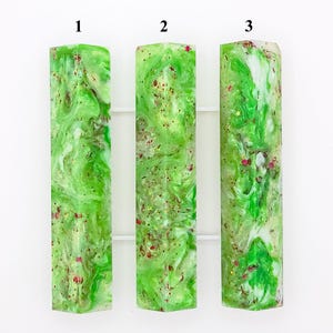 May include: Three rectangular resin blocks with a vibrant green and white swirling pattern. Each block is accented with flecks of red and gold glitter, creating a textured and decorative appearance. The blocks are displayed against a white background.