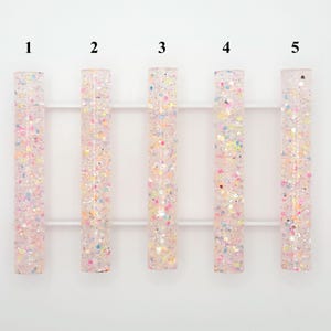 Puede incluir: Cinco piezas rectangulares transparentes de resina llenas de purpurina colorida. La purpurina incluye motas rosas, amarillas, azules y plateadas. Las piezas están dispuestas verticalmente y conectadas por una barra horizontal blanca.