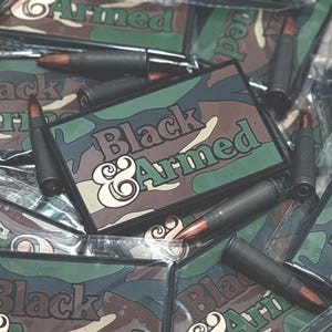 May include: A collection of rectangular patches with the text "Black & Armed" in a green and brown camouflage pattern. Several black bullets with copper tips are scattered around the patches.