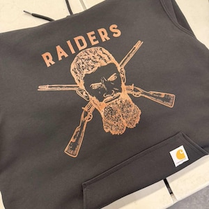 May include: Black Carhartt hoodie with a front pocket and drawstring hood. The hoodie features the word "RAIDERS" in orange lettering above an orange illustration of a bearded man with crossed rifles. A Carhartt label is on the front pocket.