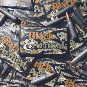 May include: A collection of rectangular patches with the words "Black & Armed" in orange and white, set against a camouflage pattern. Several bullets are scattered around the patches, creating a military-themed display.
