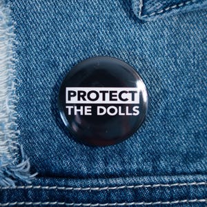 May include: A black button pin with the text "PROTECT THE DOLLS" in white, set against a denim background. The pin is round and glossy, with the text centered. The denim has visible stitching and a distressed patch.