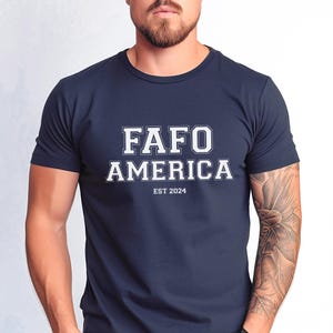 May include: Navy blue crew neck t-shirt with the white text "FAFO AMERICA" in a collegiate font. Below the text is "EST 2024". The shirt is a solid color and has short sleeves.