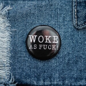 May include: A black button with white text that reads "WOKE AS FUCK!" on a blue denim background.