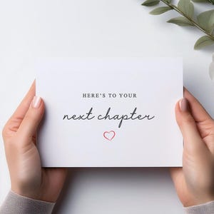 May include: A white card with the text "Here's to your next chapter" in black script font and a small red heart.