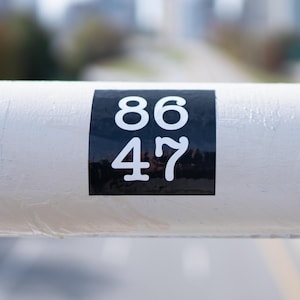 Puede incluir: Un tubo de metal blanco con una pegatina negra que dice "86 47".
