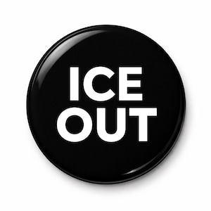 May include: A glossy black button with the words "ICE OUT" in bold white capital letters. The button is round and has a reflective surface, with the text centered on the face.