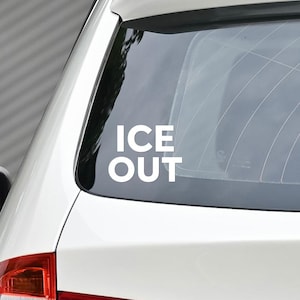 May include: White "ICE OUT" decal applied to the rear window of a white car. The text is in a bold, sans-serif font. The car's body is visible, with a portion of the roof and side panel.