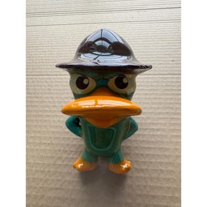 May include: A ceramic figurine of Perry the Platypus from Phineas and Ferb. The figurine is green, with an orange beak and feet, and a brown hat. The eyes are large and expressive.