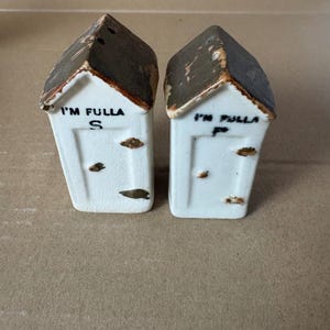 May include: A pair of vintage salt and pepper shakers shaped like small houses. The white ceramic shakers have brown roofs and the text "I'M FULLA" above the letters "S" and "P". The shakers have brown spots and a crackled finish.