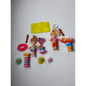 May include: A collection of two small plastic dolls with blonde hair, featuring various interchangeable outfits including skirts, tops, and pants. Accessories include two brown combs, a yellow rectangular towel, a white ring, and several small plastic bases.