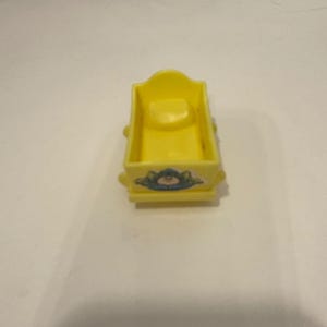 May include: A small, yellow toy cradle with a curved headboard and a rectangular base. The front of the cradle features a floral design with blue and pink accents. The cradle is made of plastic.