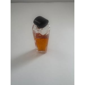 Puede incluir: Un pequeño frasco de perfume de vidrio transparente con una tapa negra. La botella está llena de un líquido marrón dorado. La botella tiene una forma geométrica única y está sobre un fondo blanco.
