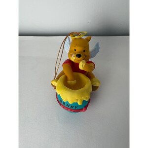 May include: A yellow bear ornament with white wings and a gold halo, wearing a red shirt. The bear sits behind a blue honey pot with yellow drips, labeled HUNNY and WAIT TILL X'MAS in black text. A gold hanging cord is attached to the top.
