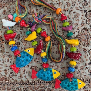 May include: A colorful handmade necklace featuring ceramic fish and various shapes in vibrant colors. The fish are blue with yellow heads and red fins. The necklace is strung on a multicolored yarn.