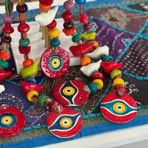 May include: Five colorful beaded necklaces with ceramic evil eye charms. The charms are red, blue, and yellow with a white eye in the center. The beads are various colors, including red, blue, green, yellow, and white.