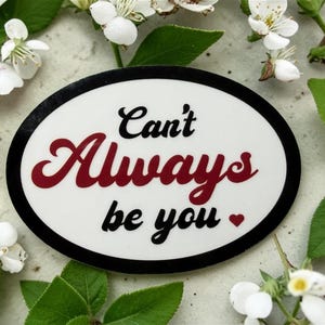 May include: An oval sticker with the text "Can't Always be you" in black and red script. The sticker is surrounded by white flowers and green leaves on a light gray surface.