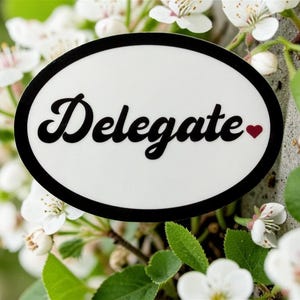May include: An oval sticker with the word "Delegate" in black script, a small red heart, and a black border. The sticker is set against a backdrop of white flowers and green leaves.