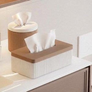 May include: A rectangular tissue box with a brown lid and a ribbed white base, alongside a cylindrical tissue holder with a brown ribbed exterior and a white lid. A bottle of perfume is also visible.