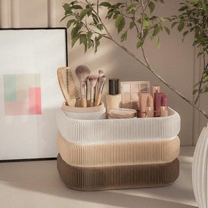 Może przedstawiać: Trzypoziomowy, żebrowany organizer do makijażu w kolorze białym, beżowym i brązowym zawiera pędzle, grzebień i różne kosmetyki. W tle znajduje się oprawione abstrakcyjne dzieło sztuki i gałąź z liśćmi.