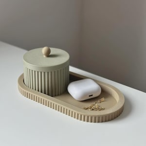 Può includere: Un vassoio decorativo verde chiaro e beige con un barattolo con coperchio abbinato e un paio di auricolari wireless bianchi. Il vassoio e il barattolo hanno un design a coste. Una collana d'oro è appoggiata sul vassoio.