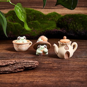 Könnte beinhalten: Drei skurrile Froschfiguren. Ein Frosch sitzt in einer Teetasse, ein anderer trägt einen Hut und hält einen kleinen Besen, und der dritte ist eine Teekanne mit einem pilzförmigen Deckel. Die Figuren sind hellgrün, beige und braun.