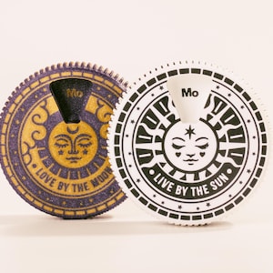 Op de afbeelding: Twee ronde, decoratieve containers met zon- en maanontwerpen. Eén is paars en goud met de tekst "LOVE BY THE MOON". De andere is zwart en wit met de tekst "LIVE BY THE SUN". Beide hebben het woord "Mo".