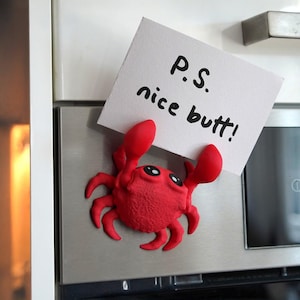 Peut inclure: Un aimant rouge en forme de crabe avec de grands yeux et des pinces, tenant une carte blanche avec le texte "P.S. nice butt!". L'aimant est fixé à un appareil en acier inoxydable.