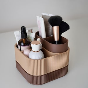 May include: A two-tiered, ribbed cosmetic organizer in shades of brown and beige. The top tier holds makeup brushes, tubes, and bottles. The organizer is designed to hold beauty products and is ideal for a vanity or bathroom.
