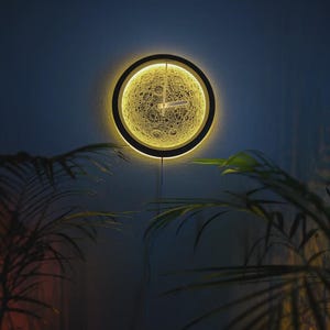 May include: A round wall clock with a yellow moon-like face and black frame. The clock hands are visible against the illuminated face, and the clock is mounted on a dark blue wall. The clock is surrounded by green leafy plants.