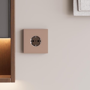 Peut inclure: Une prise électrique murale carrée de couleur rose poudré mat avec un centre circulaire. La prise présente deux trous ronds pour les broches et deux clips de mise à la terre métalliques sur les côtés, montée à fleur sur un mur lisse et clair.