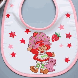 May include: White baby bib with a pink trim and a Strawberry Shortcake design. The bib features a cartoon Strawberry Shortcake character, a pink cat, red and pink stars, and a strawberry.