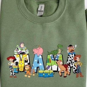 Op de afbeelding: Saliegroene sweatshirt met het woord "MAMA" en personages uit een populaire animatiefilm. De letters zijn versierd met afbeeldingen van Buzz Lightyear, Hamm, Rex, Mr. Potato Head en anderen. Een leuk, kleurrijk ontwerp.