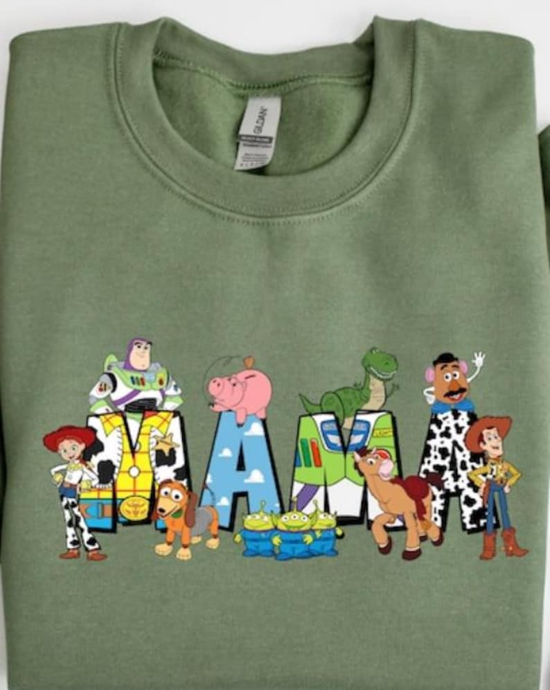 Op de afbeelding: Saliegroene sweatshirt met het woord "MAMA" en personages uit een populaire animatiefilm. De letters zijn versierd met afbeeldingen van Buzz Lightyear, Hamm, Rex, Mr. Potato Head en anderen. Een leuk, kleurrijk ontwerp.