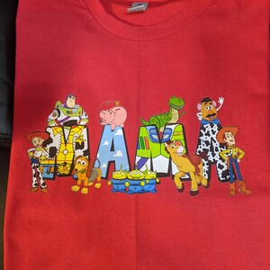 Op de afbeelding: Rood T-shirt met het woord "MAMA" in een ontwerp met personages uit de film Toy Story. Het ontwerp bevat Woody, Buzz Lightyear en andere personages.