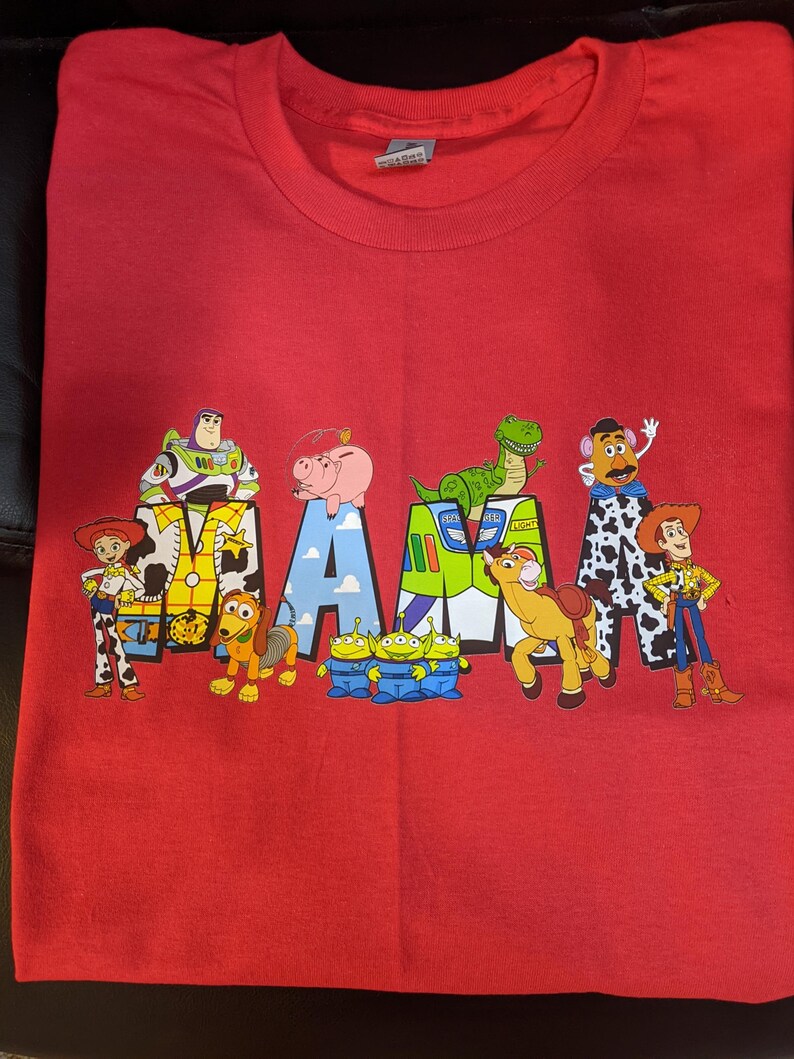 Op de afbeelding: Rood T-shirt met het woord "MAMA" in een ontwerp met personages uit de film Toy Story. Het ontwerp bevat Woody, Buzz Lightyear en andere personages.