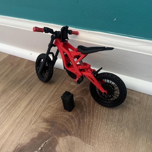 以下が含まれることがあります： 黒いシートと車輪が付いた赤と黒のミニチュア電動バイク。 モデルは木製の表面に置かれ、隣には小さな黒い箱があります。 ハンドルバーとアクセントは赤です。