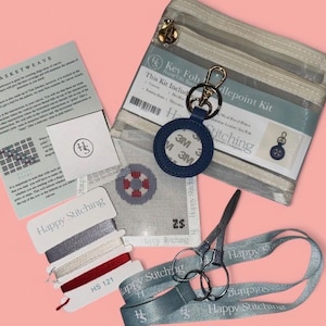 Puede incluir: Un kit de punto de aguja con un llavero de cuero azul marino, tijeras, hilo y un lienzo con un diseño rojo y gris. El kit incluye una bolsa con cremallera transparente e instrucciones. El texto "Happy Stitching" es visible.