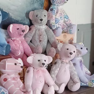 May include: A collection of handmade teddy bears in various pastel colors. The bears feature unique patterns, including hearts and stripes, and are adorned with decorative details like buttons and frills. The bears are arranged on a wooden surface.