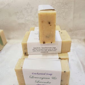 May include: A stack of handmade soap bars with a white label that reads "Enchanted Soap Lemongrass & Lavender Olive Oil Soap with Goats Milk & Shea Butter www.hesoaps.com".