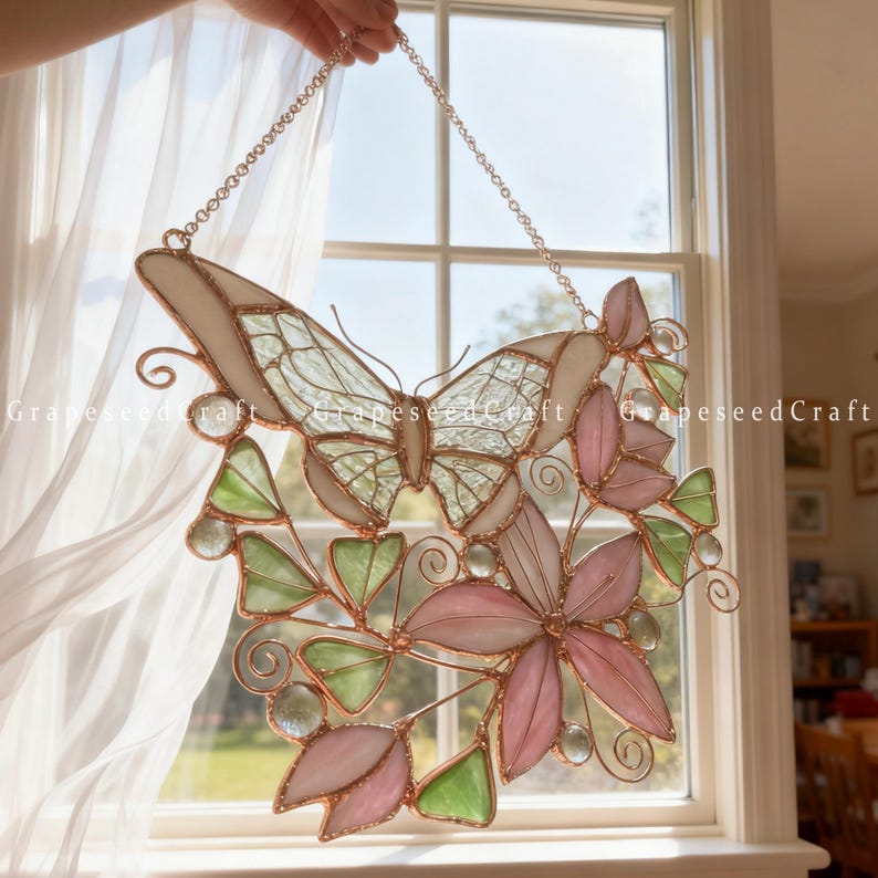 May include: A stained glass butterfly and floral window hanging. The butterfly has a cream-colored body and wings with a clear, textured pattern. Pink and green flowers and leaves are arranged below the butterfly, all framed with copper wire.
