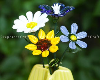 May include: A bouquet of colorful glass flowers in a yellow vase. The arrangement includes a white daisy, a yellow sunflower, a blue flower, and a light blue flower. The flowers are on green stems.