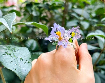 May include: A handcrafted glass ring with a translucent purple flower design, featuring yellow and green details. The ring is held up against a backdrop of green foliage. The text "GrapeseedCraft" is visible on the side.