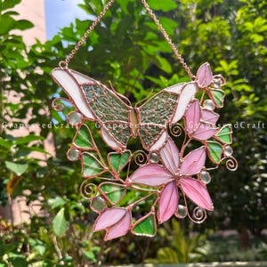 May include: Stained glass butterfly and flower suncatcher with pink, green, and white glass. The butterfly has a green textured center and white wings. Pink flowers and green leaves surround the butterfly. The suncatcher is suspended by a copper chain.