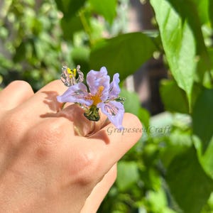 Puede incluir: Un anillo artesanal con una delicada flor de iris de color lila claro con detalles en amarillo y verde. El anillo es de vidrio y tiene una banda plateada. El texto "GrapeSeed Craft" es visible.