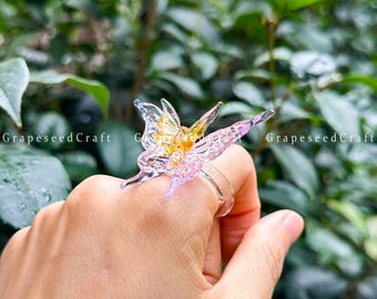 May include: A clear resin butterfly ring with yellow, pink, and purple accents. The ring is held up by a hand in front of a green leafy background. The text "GrapeseedCraft" is visible.