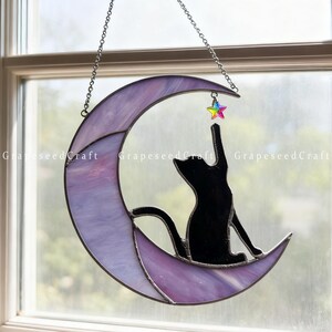 May include: A stained glass crescent moon with a black cat silhouette reaching for a colorful star. The moon is purple and white, and the star is multi-colored. The piece is suspended by a chain.