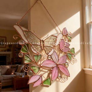 May include: Stained glass butterfly and floral wall hanging. The artwork features a butterfly with white and tan wings, and pink and green flowers. The piece is framed with copper-colored metal and hangs from a chain.