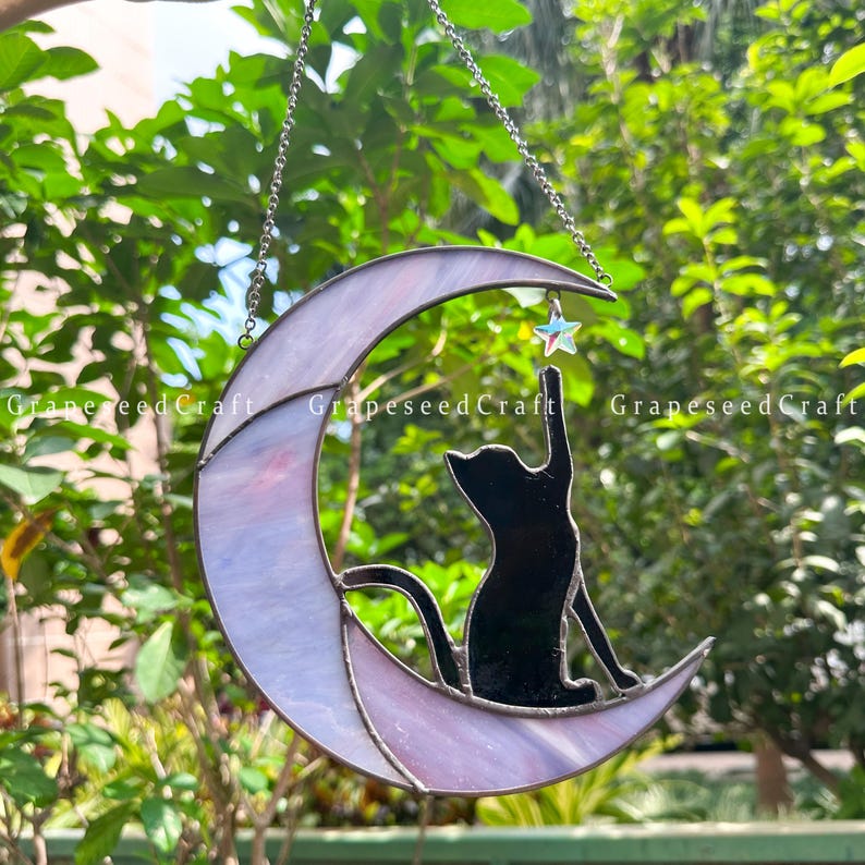May include: Stained glass crescent moon with a black cat silhouette reaching for a star. The moon is a blend of lavender and white, with a silver chain for hanging. A charming cat-themed decoration.
