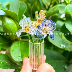 Può includere: Un piccolo vaso di vetro con un bouquet di fiori di vetro colorati. I fiori sono in tonalità di blu, giallo e trasparente, con dettagli delicati. Il vaso è trasparente con scanalature verticali. Sfondo verde sfocato.